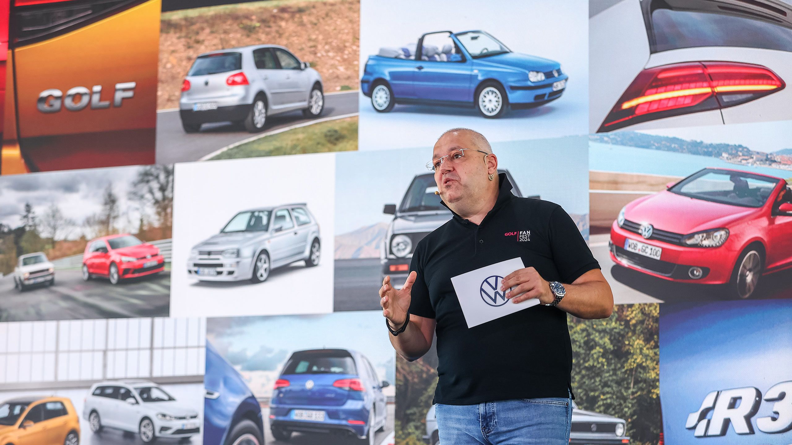 Volkswagen Tech Talks A man speaks at a presentation holding a Volkswagen logo card.