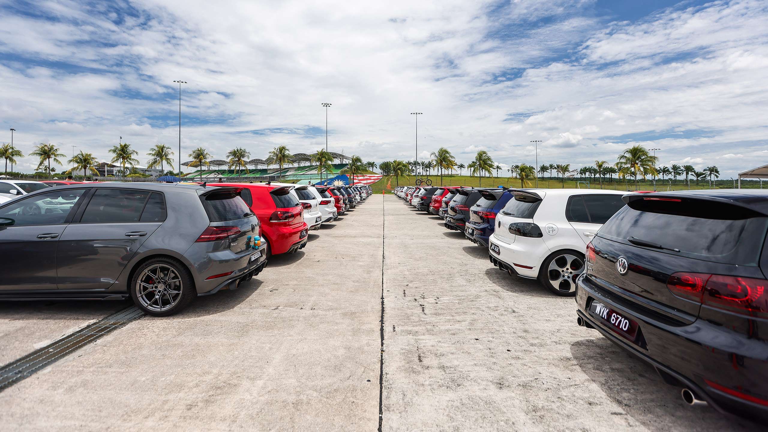 Golf Gathering A parking lot filled with various colored cars, mostly hatchbacks. Palm trees are visible in the background.