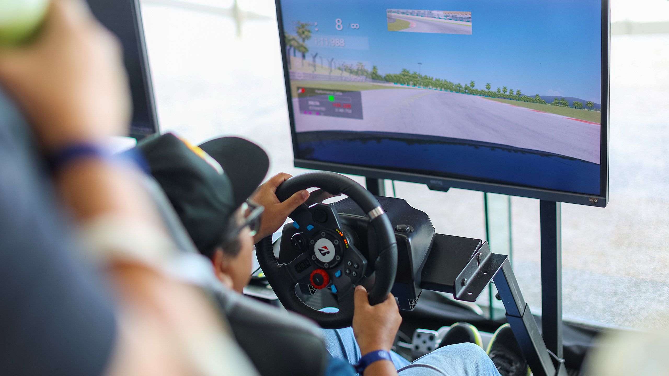 Racing simulator Person using a racing simulator setup with a steering wheel and pedals, driving on a virtual track displayed on a screen.