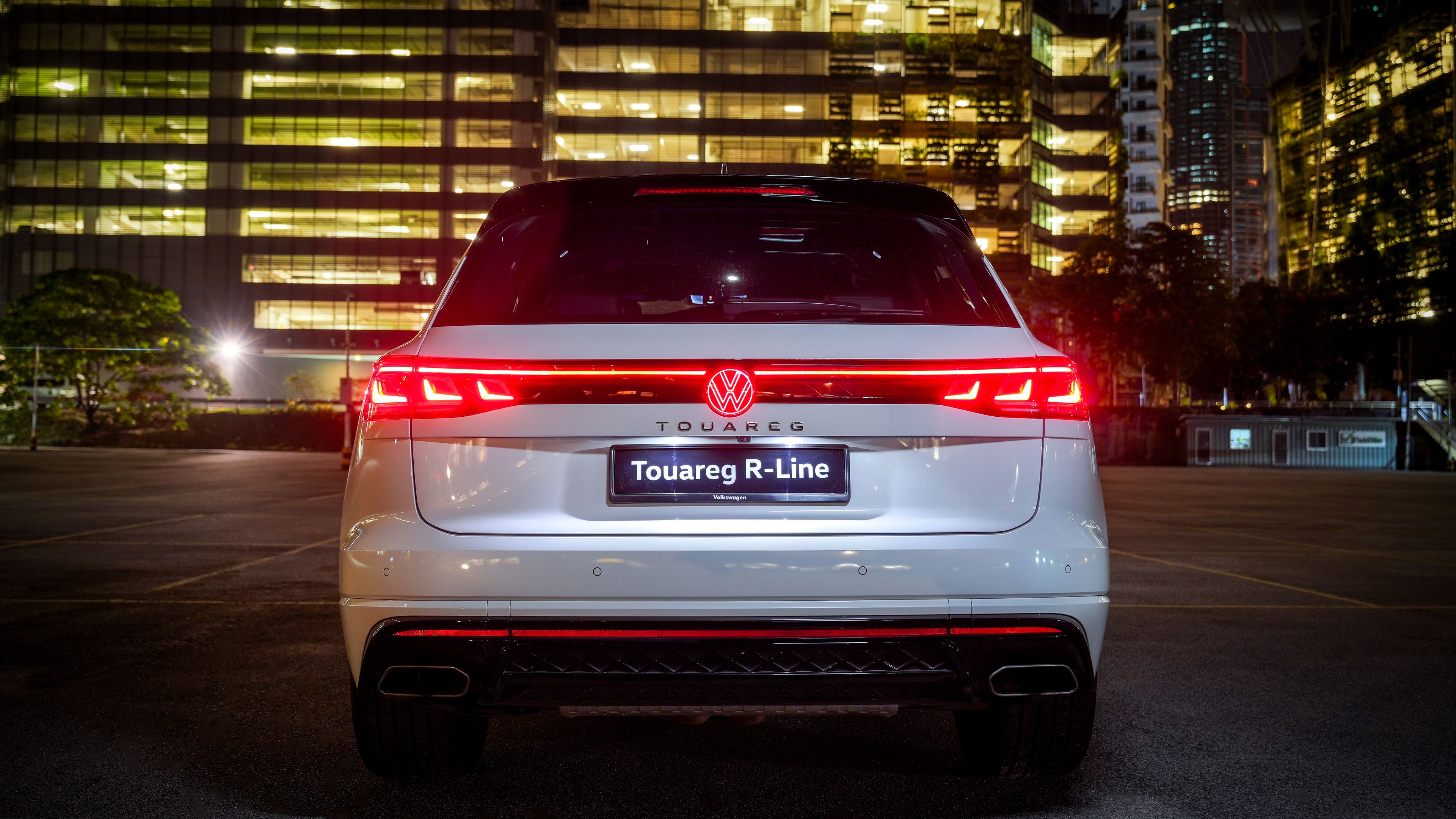 Volkswagen Touareg R-Line Rear view of a Volkswagen Touareg R-Line luxury SUV with lit tail lights at night, set against an urban backdrop.