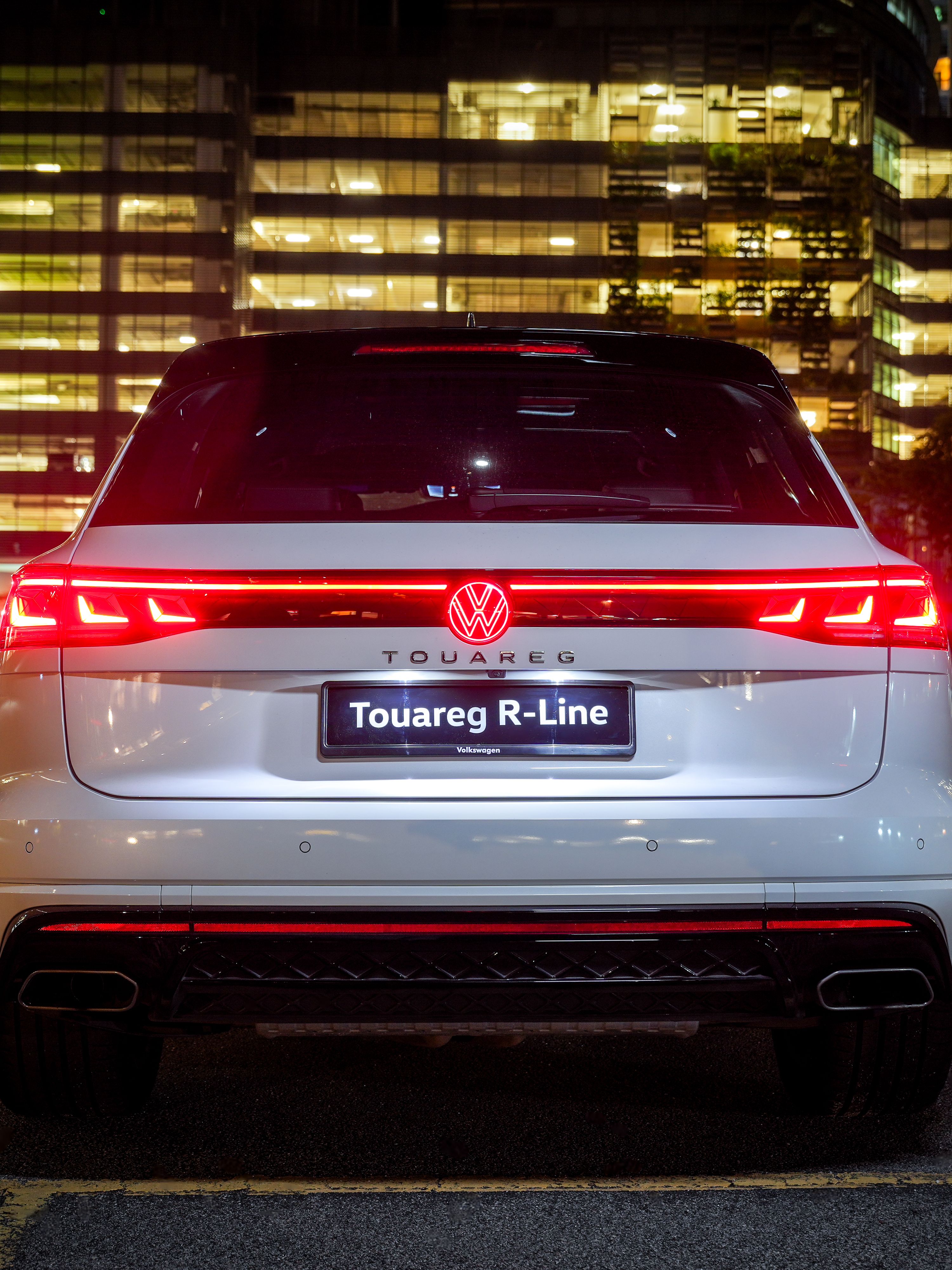 Volkswagen Touareg R-Line Rear view of a Volkswagen Touareg R-Line luxury SUV with lit tail lights at night, set against an urban backdrop.