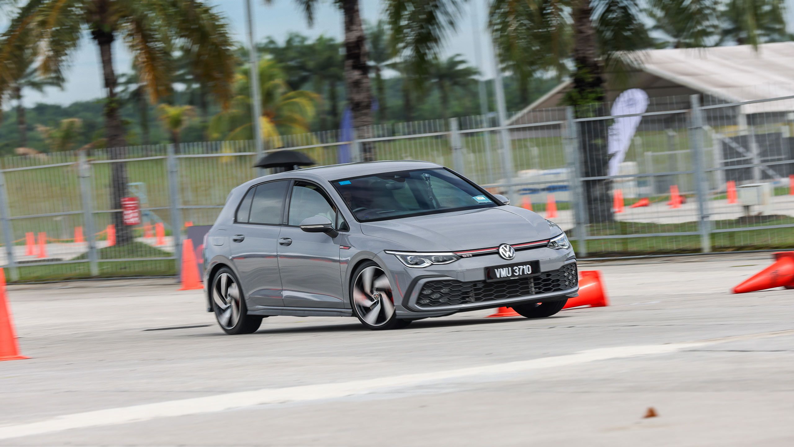 Golf Gymkhana A gray Volkswagen hatchback performs a driving maneuver around orange traffic cones on a paved area.
