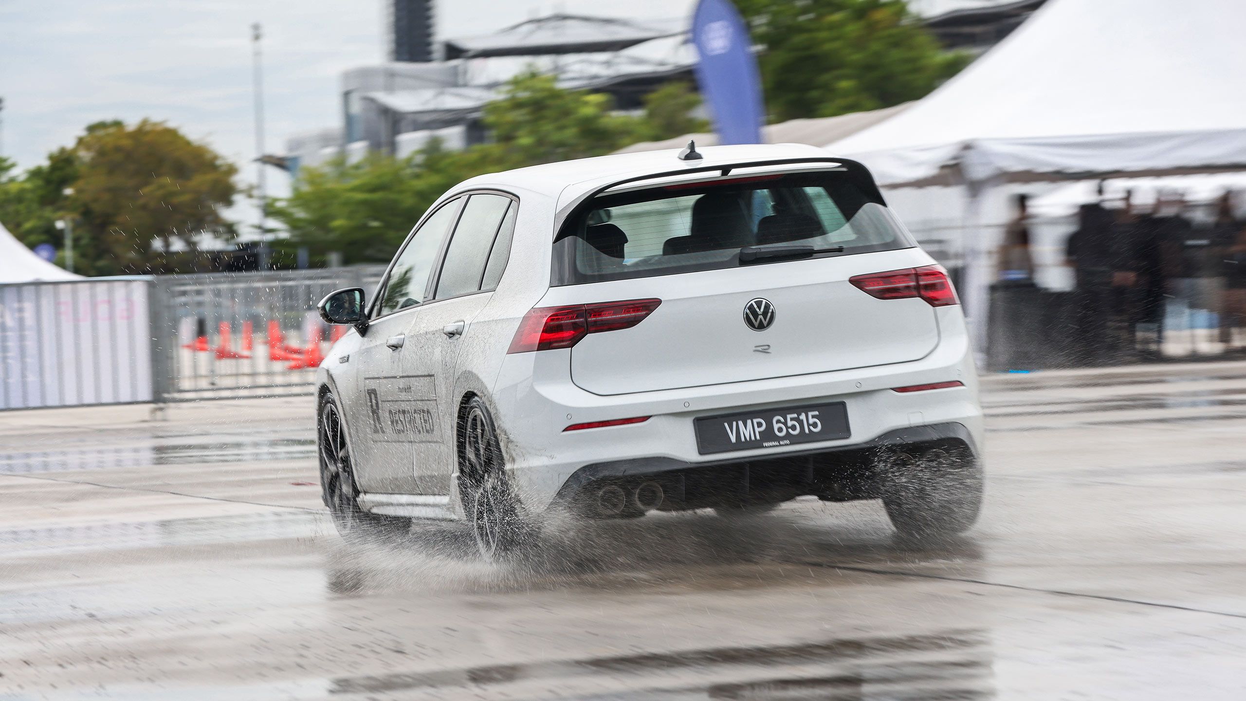 Drift Taxi A white Volkswagen car drives on a wet surface at an outdoor event, creating splashback from the water on the ground.